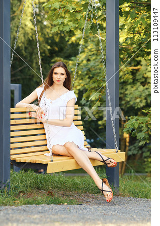 Cheerful woman in white dress enjoys swinging on a swing in city park 113109447