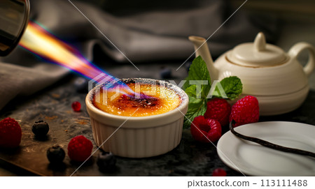 The chef burns sugar on a portion of creme brulee, the dessert is served with fresh berries and mint leaves, a popular dessert made from cream 113111488