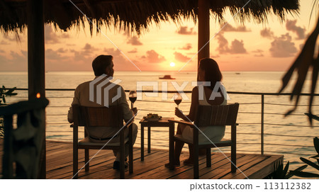 An adult married couple at a table in a restaurant with glasses of red wine enjoys a conversation and admires the beautiful sunsets in the sea, advertising a restaurant or cafe An adult married couple at a table in a restaurant with glasses of red wine enjoys a conversation and admires the beautiful sunsets in the sea, advertising a restaurant or cafe 113112382
