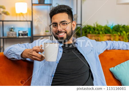 Tired smiling Indian man drinking cup of warm coffee or herbal tea in the morning relaxing enjoying 113116081
