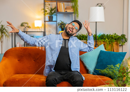 Energetic young Indian man singing dancing with hands sitting on couch room enjoying listening music Energetic young Indian man singing dancing with hands sitting on couch room enjoying listening music 113116122