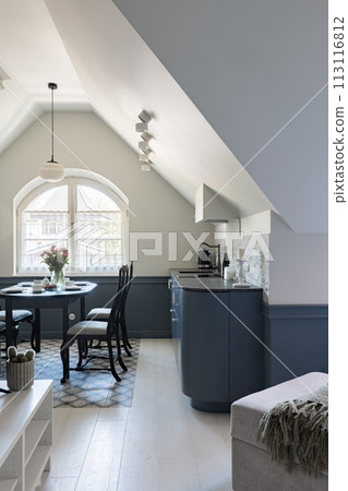 A kitchen in a building with table and chairs, lit by a fixture near the window 113116812