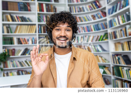 Cheerful student engaging in online learning, making a friendly gesture to the camera while using a headset in a library. Cheerful student engaging in online learning, making a friendly gesture to the camera while using a headset in a library. 113116960
