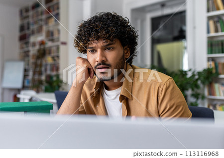 Young adult with curly hair appears serious and focused while working on a laptop in a bright, contemporary office space. Young adult with curly hair appears serious and focused while working on a laptop in a bright, contemporary office space. 113116968