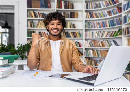 Joyful young man triumphantly clenches fist looking at laptop in library, expressing excitement for his achievement or win. Joyful young man triumphantly clenches fist looking at laptop in library, expressing excitement for his achievement or win. 113116970