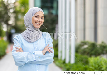 Portrait of a happy and confident Muslim woman wearing a hijab, smiling warmly with arms crossed in an urban park setting. Portrait of a happy and confident Muslim woman wearing a hijab, smiling warmly with arms crossed in an urban park setting. 113121196