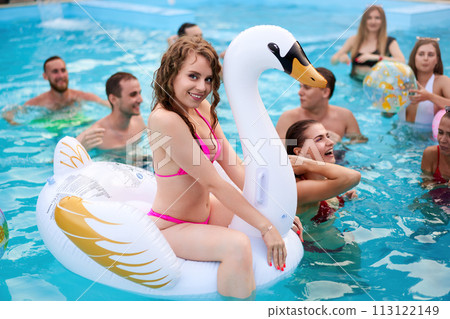 Pretty woman in bikini swimsuit hanging out on inflatable white swan mattress on night pool party. Friends partying with cocktails in holiday villa. 113122149