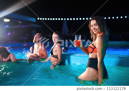Pretty woman in bikini drinking cocktail and hanging out with friends at night pool party. People having fun dancing with drinks and splashing water in luxury private villa swimming pool. Pretty woman in bikini drinking cocktail and hanging out with friends at night pool party. People having fun dancing with drinks and splashing water in luxury private villa swimming pool. 113122176