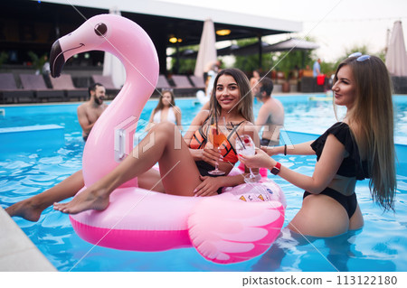 Pretty woman in bikini swimwear hanging out on inflatable pink flamingo mattress on night pool party. Friends partying with cocktails in resort villa swimming pool. Girls toast, cheer with drinks. Pretty woman in bikini swimwear hanging out on inflatable pink flamingo mattress on night pool party. Friends partying with cocktails in resort villa swimming pool. Girls toast, cheer with drinks. 113122180