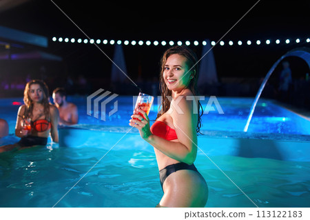 Pretty woman in bikini drinking cocktail and hanging out with friends at night pool party. People having fun dancing with drinks and splashing water in luxury private villa swimming pool. Pretty woman in bikini drinking cocktail and hanging out with friends at night pool party. People having fun dancing with drinks and splashing water in luxury private villa swimming pool. 113122183