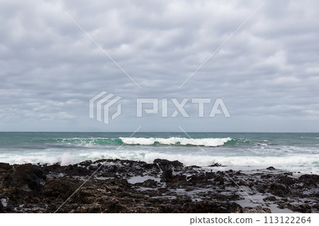Surf on a rocky coast in front of the horizon under a cloudy sky. Tropical stormy ocean with waves crashing on the rocks. Stormy sea with stormy clouds and black lava rocks on the shore. Splashing 113122264
