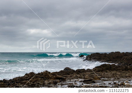 Surf on a rocky coast in front of the horizon under a cloudy sky. Tropical stormy ocean with waves crashing on the rocks. Stormy sea with stormy clouds and black lava rocks on the shore. Splashing 113122275