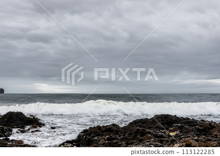Surf on a rocky coast in front of the horizon under a cloudy sky. Tropical stormy ocean with waves crashing on the rocks. Stormy sea with stormy clouds and black lava rocks on the shore. Splashing Surf on a rocky coast in front of the horizon under a cloudy sky. Tropical stormy ocean with waves crashing on the rocks. Stormy sea with stormy clouds and black lava rocks on the shore. Splashing 113122285