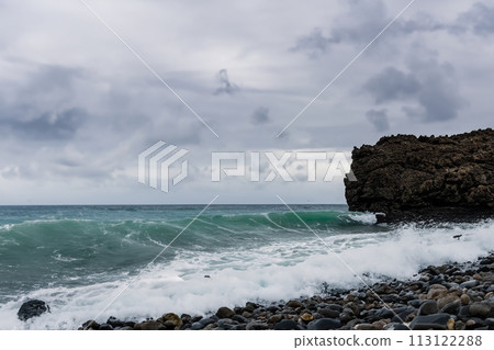 Surf on a rocky coast in front of the horizon under a cloudy sky. Tropical stormy ocean with waves crashing on the rocks. Stormy sea with stormy clouds and black lava rocks on the shore. Splashing 113122288