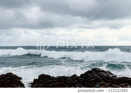 Surf on a rocky coast in front of the horizon under a cloudy sky. Tropical stormy ocean with waves crashing on the rocks. Stormy sea with stormy clouds and black lava rocks on the shore. Splashing Surf on a rocky coast in front of the horizon under a cloudy sky. Tropical stormy ocean with waves crashing on the rocks. Stormy sea with stormy clouds and black lava rocks on the shore. Splashing 113122305