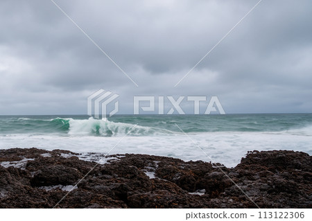Surf on a rocky coast in front of the horizon under a cloudy sky. Tropical stormy ocean with waves crashing on the rocks. Stormy sea with stormy clouds and black lava rocks on the shore. Splashing Surf on a rocky coast in front of the horizon under a cloudy sky. Tropical stormy ocean with waves crashing on the rocks. Stormy sea with stormy clouds and black lava rocks on the shore. Splashing 113122306