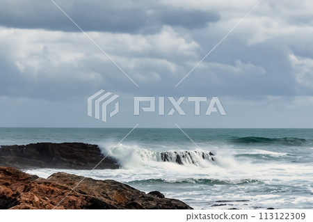 Surf on a rocky coast in front of the horizon under a cloudy sky. Tropical stormy ocean with waves crashing on the rocks. Stormy sea with stormy clouds and black lava rocks on the shore. Splashing Surf on a rocky coast in front of the horizon under a cloudy sky. Tropical stormy ocean with waves crashing on the rocks. Stormy sea with stormy clouds and black lava rocks on the shore. Splashing 113122309