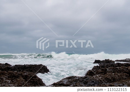 Surf on a rocky coast in front of the horizon under a cloudy sky. Tropical stormy ocean with waves crashing on the rocks. Stormy sea with stormy clouds and black lava rocks on the shore. Splashing 113122315