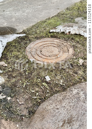 A rusty iron hatch with a pattern is located next to a dry grass pedestrian road during the daytime. Close-up 113124318