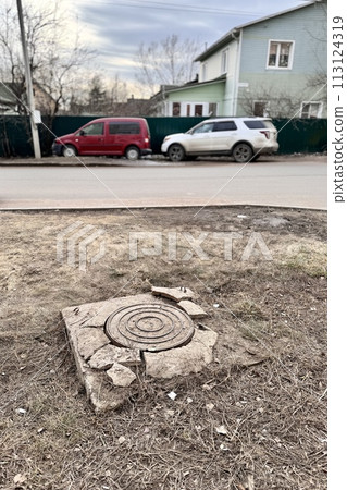 A rusty iron hatch with a pattern and a large gap at the top is mounted in a broken cracked asphalt structure, a well next to the road and an apartment building with two passenger cars 113124319