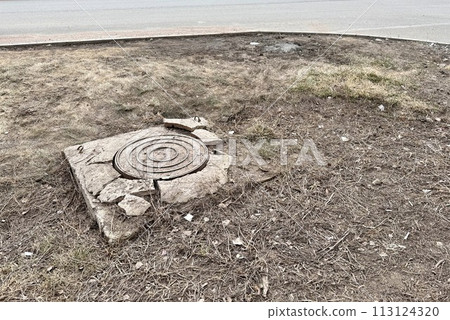 A rusty iron hatch with a pattern and a large gap at the top is mounted in a broken cracked asphalt structure well next to the road. Close-up A rusty iron hatch with a pattern and a large gap at the top is mounted in a broken cracked asphalt structure well next to the road. Close-up 113124320