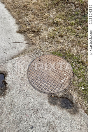 A rusty iron hatch with a pattern with a large gap at the top is mounted in the asphalt pavement of the pedestrian road. Close-up 113124352