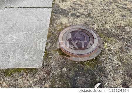 A rusty iron hatch with a pattern is located next to a dry grass pedestrian road during the daytime. Close-up 113124354