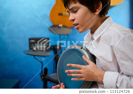 Close-up portrait teen boy with eyes closed, playing the djembe drum in a cozy home music studio, banging drums, creating the rhythm of ethnic music. Authentic African percussion musical instrument Close-up portrait teen boy with eyes closed, playing the djembe drum in a cozy home music studio, banging drums, creating the rhythm of ethnic music. Authentic African percussion musical instrument 113124726