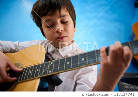 Close-up portrait of confident teenager boy plucking strings while strumming his guitar with delight. Inspired teenager boy playing the acoustic guitar Close-up portrait of confident teenager boy plucking strings while strumming his guitar with delight. Inspired teenager boy playing the acoustic guitar 113124741
