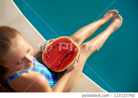 Prety blonde girl holding half of watermelon in the blue pool on slim legs. Young woman wearing blue stripedbody swimsuit. Instagram style. Tropical fruit diet. Summer holiday idyllic concept. Prety blonde girl holding half of watermelon in the blue pool on slim legs. Young woman wearing blue stripedbody swimsuit. Instagram style. Tropical fruit diet. Summer holiday idyllic concept. 113125760