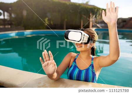 Young woman in bikini swimsuit using waterproof virtual reality glasses in swimming pool playing game. Blonde girl with vr headset touches objects with her hands. VR technology and vacation concept. 113125780