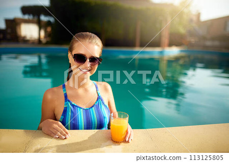 Young woman relaxing in a pool in summer with glass of orange juice. Pretty caucasian blonde girl wearing blue swimsuit and sunglasses. Travel and vacation concept. 113125805