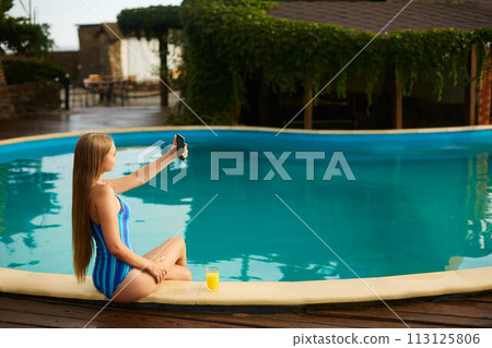 Attractive blonde woman with long hair sitting at poolside and taking selfie portrait with cellphone camera. Slim girl wear blue striped body swimsuit and holds a glass with orange juice. Attractive blonde woman with long hair sitting at poolside and taking selfie portrait with cellphone camera. Slim girl wear blue striped body swimsuit and holds a glass with orange juice. 113125806