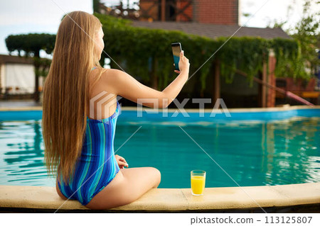 Young woman relaxing in a pool in summer with glass of orange juice. Pretty caucasian blonde girl wearing blue swimsuit. Travel and vacation concept. Young woman relaxing in a pool in summer with glass of orange juice. Pretty caucasian blonde girl wearing blue swimsuit. Travel and vacation concept. 113125807
