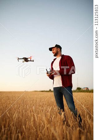 Farmer holds remote controller with his hands while quadcopter is flying on background. Drone hovers behind the agronomist in wheat field. Agricultural new technologies and innovations. Back view Farmer holds remote controller with his hands while quadcopter is flying on background. Drone hovers behind the agronomist in wheat field. Agricultural new technologies and innovations. Back view 113125813