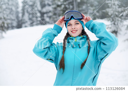 Smiling nordic woman with pigtails puts on protective ski goggles. Snowboarder girl touching mask at ski resort on snowfall near forest. Blue eyed elegant sportswoman in colorful fashionable outfit. Smiling nordic woman with pigtails puts on protective ski goggles. Snowboarder girl touching mask at ski resort on snowfall near forest. Blue eyed elegant sportswoman in colorful fashionable outfit. 113126084