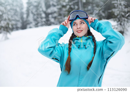 Smiling nordic woman with pigtails puts on protective ski goggles. Snowboarder girl touching mask at ski resort on snowfall near forest. Blue eyed elegant sportswoman in colorful fashionable outfit. 113126090