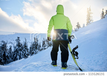 Back view of snowboarder climbing with his board on the mount for backcountry freeride session in the forest on sunset. Man with snowboard walking at ski resort. Rider lime fashionable outfit. Back view of snowboarder climbing with his board on the mount for backcountry freeride session in the forest on sunset. Man with snowboard walking at ski resort. Rider lime fashionable outfit. 113126131