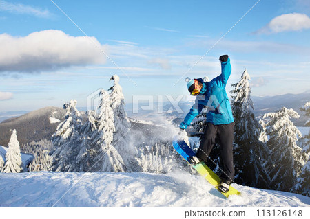 Snowboarder doing tricks at ski resort. Rider performing jump with his snowboard near forest on backcountry freeride or freestyle session in colorful fashionable outfit. Copyspace area. Powder in air. 113126148