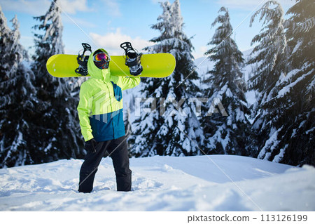 Snowboarder climbing to mountain top carrying his snowboard through forest for backcountry freeride and wearing reflective goggles, colorful fashion outfit and balaclava at ski resort. Winter sports. Snowboarder climbing to mountain top carrying his snowboard through forest for backcountry freeride and wearing reflective goggles, colorful fashion outfit and balaclava at ski resort. Winter sports. 113126199