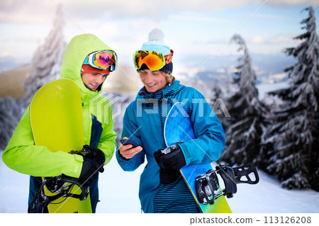 Two snowboarders talking and holding smartphones. Friends looking at cellphone and communicating in social network sharing their photos in app with internet connection wearing colorful fashion outfit. 113126208