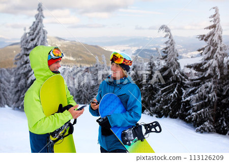Two snowboarders talking and holding smartphones. Friends looking at cellphone and communicating in social network sharing their photos in app with internet connection wearing colorful fashion outfit. Two snowboarders talking and holding smartphones. Friends looking at cellphone and communicating in social network sharing their photos in app with internet connection wearing colorful fashion outfit. 113126209
