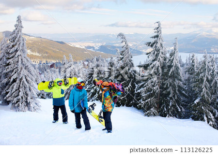 Three snowboarders walking at ski resort. Friends climbing to mountain top carrying their snowboards through forest for backcountry freeride and wearing reflective goggles, colorful fashion clothes. Three snowboarders walking at ski resort. Friends climbing to mountain top carrying their snowboards through forest for backcountry freeride and wearing reflective goggles, colorful fashion clothes. 113126225