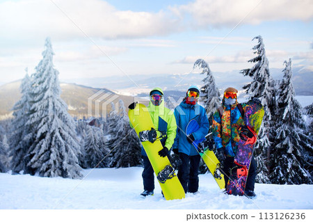 Three snowboarders posing at ski resort. Friends climbing to mountain top carrying their snowboards through forest for backcountry freeride and wearing reflective goggles, colorful fashion clothes. Three snowboarders posing at ski resort. Friends climbing to mountain top carrying their snowboards through forest for backcountry freeride and wearing reflective goggles, colorful fashion clothes. 113126236