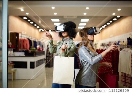Two women in modern virtual reality headsets having expirience in shopping at lingerie store. Multiracial girls in vr glasses with bags touching and pointing interface elements in underwear shop. 113126309