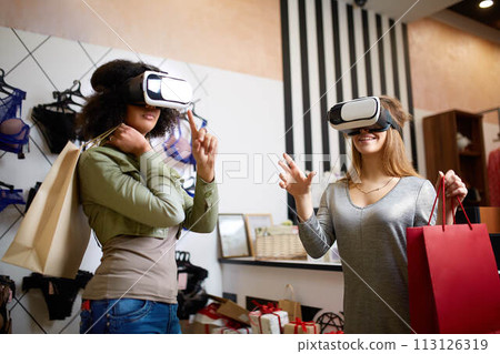 Two women in modern virtual reality headsets having expirience in shopping at lingerie store. Multiracial girls in vr glasses with bags touching and pointing interface elements in underwear shop. 113126319
