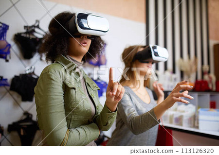 Two women in modern virtual reality headsets having expirience in shopping at lingerie store. Multiracial girls in vr glasses with bags touching and pointing interface elements in underwear shop. 113126320