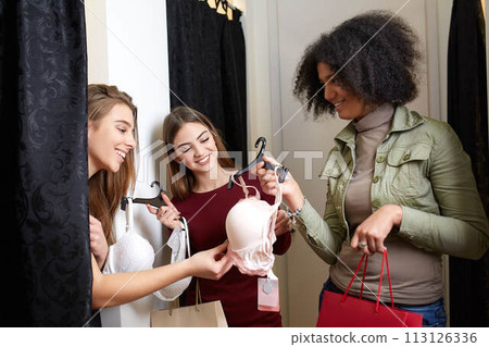 Beautiful young woman asking for a friends opinion in the fitting room of lingerie store. Female tries on bra in underwear shop. Shop assistant helps girl to choose right size in changing room. 113126336