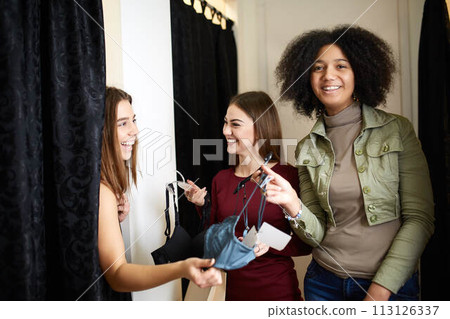 Beautiful young woman asking for a friends opinion in the fitting room of lingerie store. Female tries on bra in underwear shop. Shop assistant helps girl to choose right size in changing room. 113126337