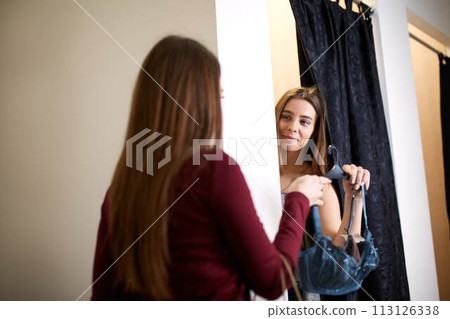 Beautiful young woman asking for a friends opinion in the fitting room of lingerie store. Female tries on bra in underwear shop. Shop assistant helps girl to choose right size in changing room. 113126338